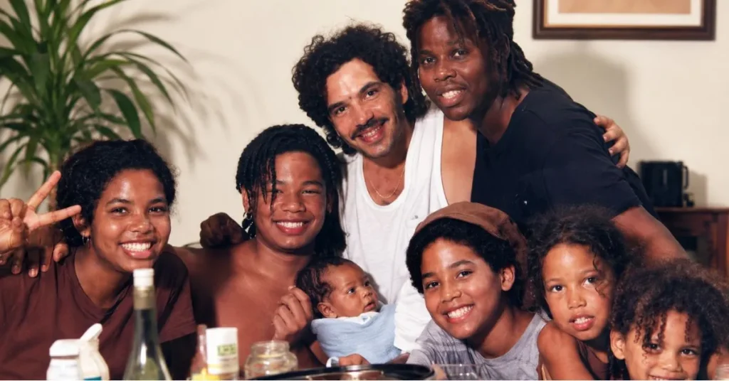 A smiling family group portrait indoors, featuring adults, teenagers, and children gathered closely together around a dining table in a warm and cozy setting.