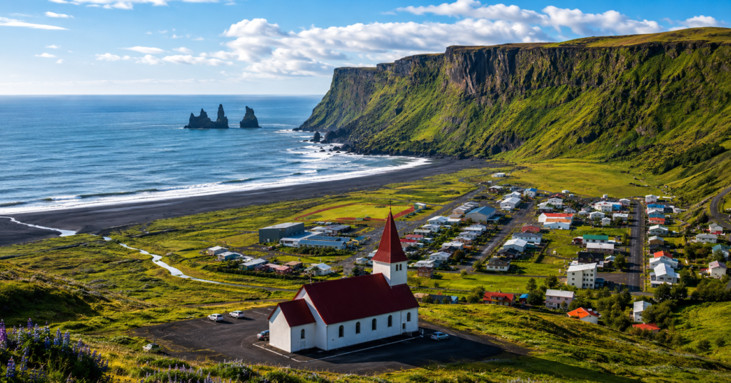 Panoramic view of Vik village Iceland with black sand coastline, green mountains, and iconic red-roof church under bright blue skies