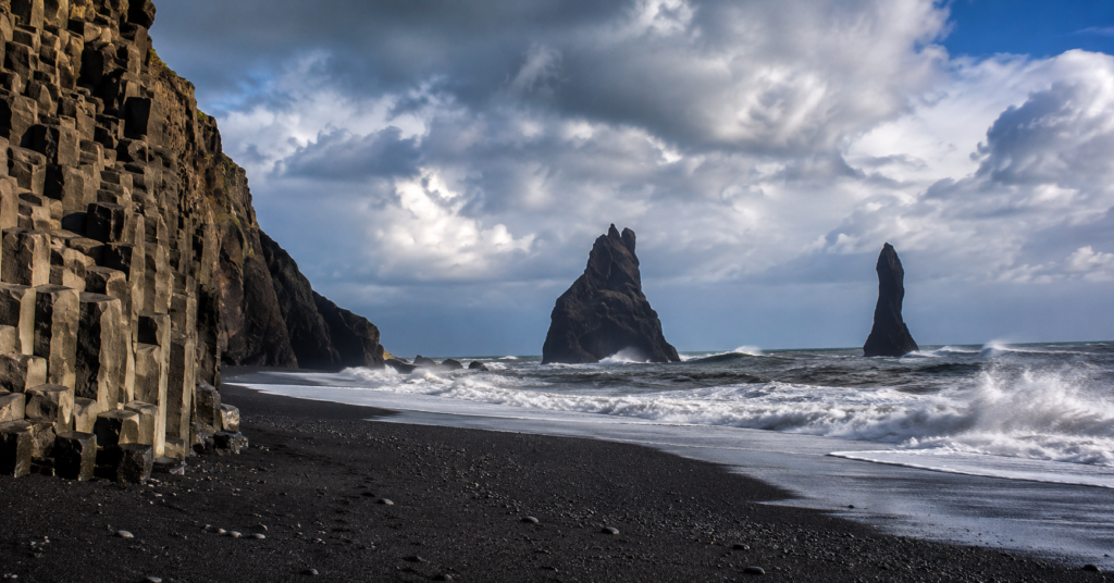 Reynisfjara black sand beach in Vik Iceland with basalt cliffs, crashing Atlantic waves, and Reynisdrangar sea stacks under dramatic skies