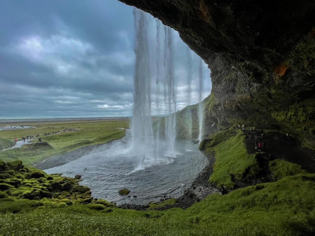 View from behind Seljalandsfoss waterfall in Iceland with cascading water, moss-covered cliffs, and dramatic cloudy skies over the green landscape.