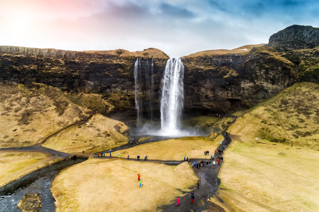 Wide landscape view of Seljalandsfoss waterfall in Iceland with visitors walking along scenic paths surrounded by rugged cliffs and open grassy terrain.