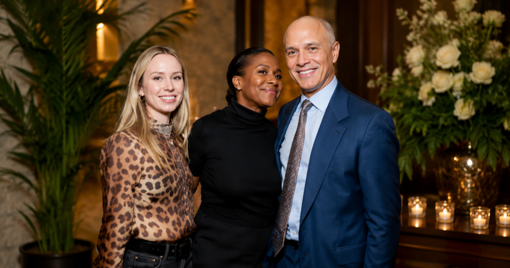 Emily Threlkeld posing with guests at an elegant indoor event, wearing a stylish leopard-print blouse beside a well-dressed couple in a luxury floral setting.