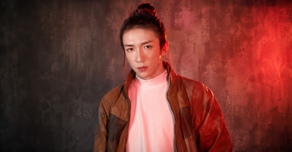 Dancer Mo poses in a stylish portrait wearing a brown leather jacket over a white high-neck top, standing against a vibrant red-lit backdrop with a confident expression.