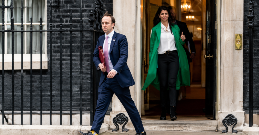 Gina Coladangelo in a green coat exits a government-style building doorway while a man in a navy suit carrying a folder walks past in the foreground.