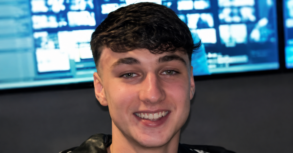 Jay Slater smiling in an indoor portrait with short dark hair, photographed against a background of illuminated screens.