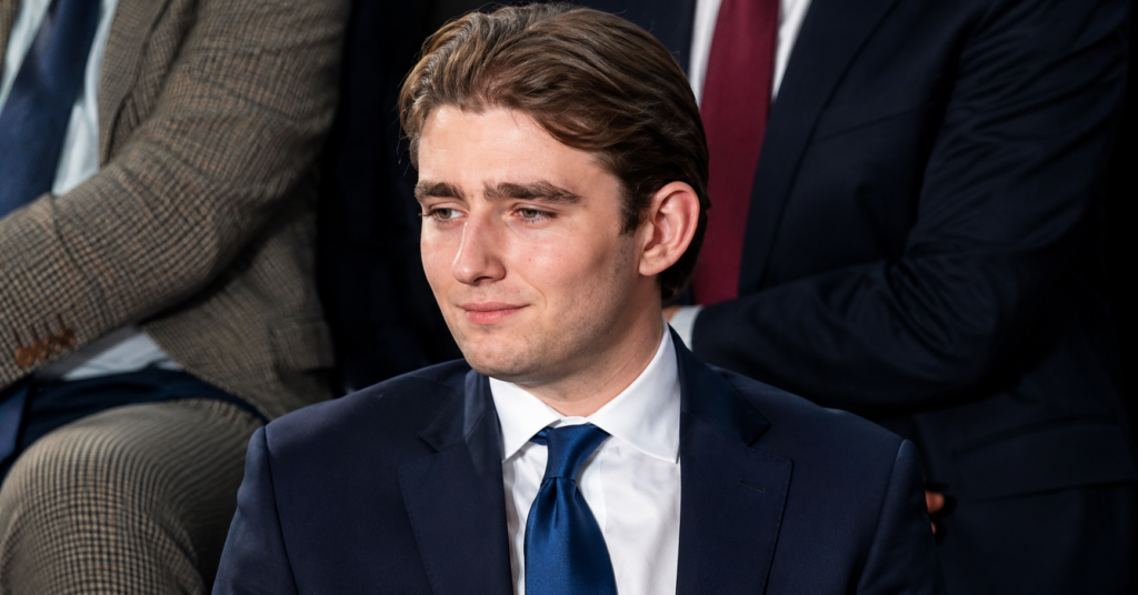 Barron Trump seated in a navy blue suit and blue tie during a formal public event, captured in a high-resolution editorial-style portrait with a blurred audience in the background.