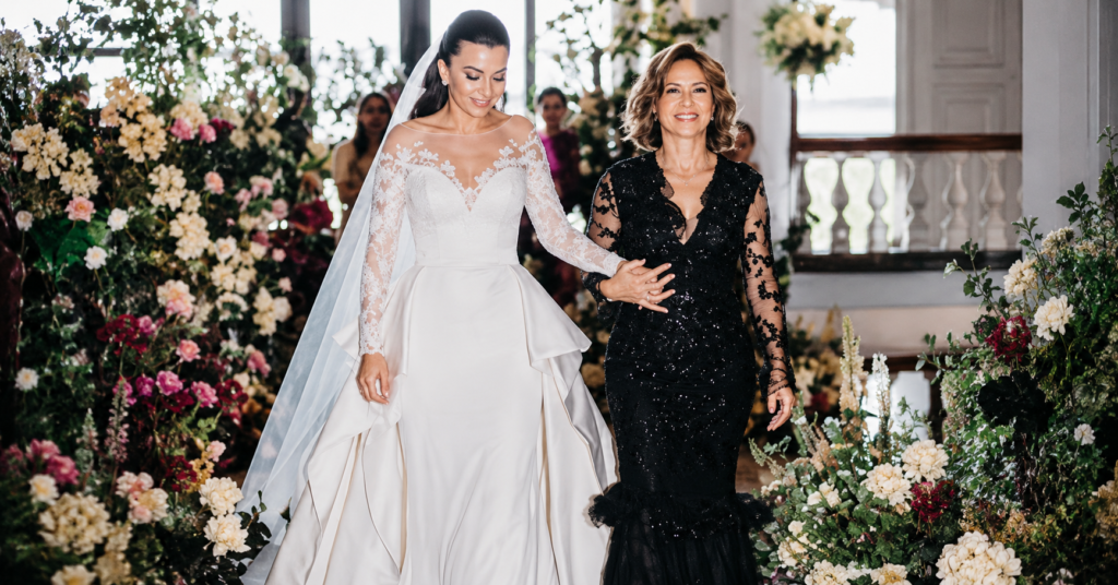 Deshanna Marie Minuto wedding moment as she walks down a flower-lined aisle in an elegant white gown alongside a family member during a beautiful ceremony.