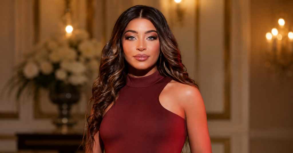 Gia Giudice posing in an elegant burgundy dress with long wavy hair, photographed in a glamorous indoor setting with warm lighting.