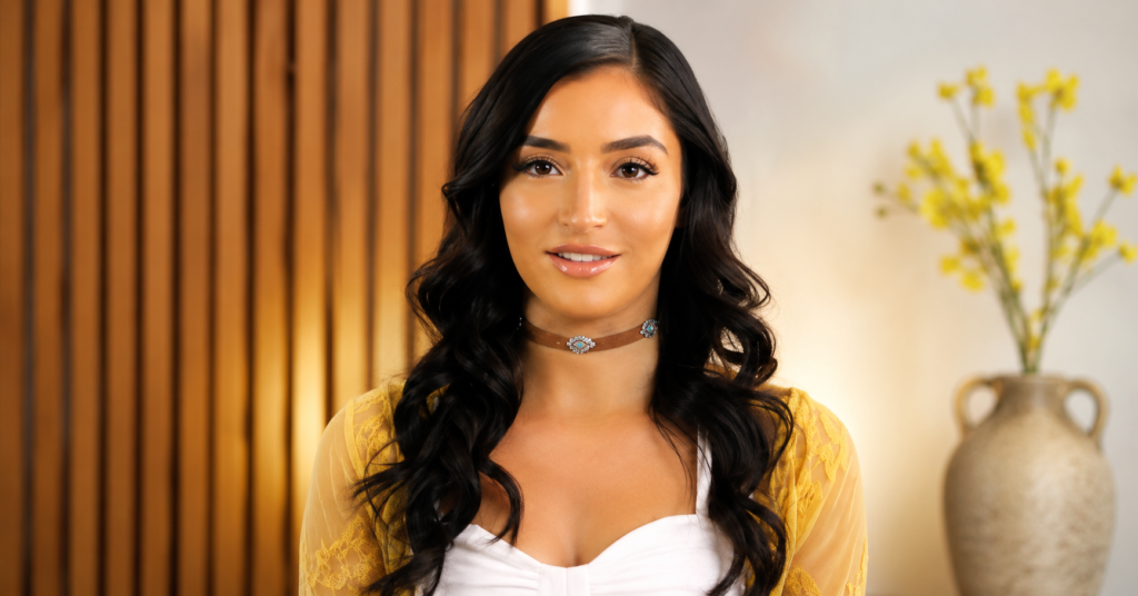 Emily Willis with long dark wavy hair wearing a white top and yellow cardigan, smiling at the camera.