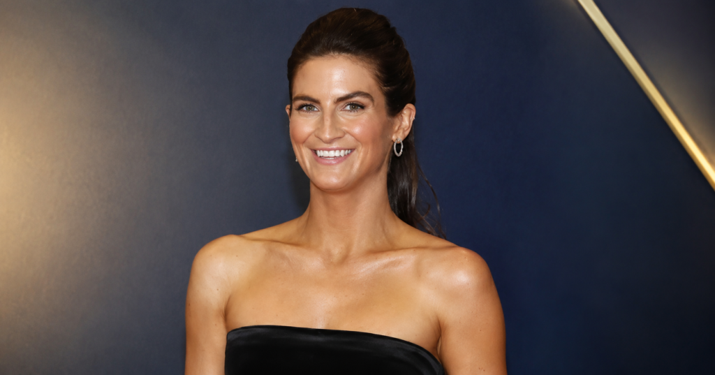 Kaitlan Collins smiling in a strapless black dress at a media event with a blue backdrop.