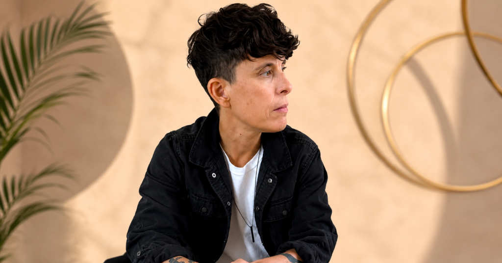 Portrait of Andrea Gibson wearing a black jacket over a white T-shirt, looking to the side with short curly hair and visible forearm tattoos against a soft neutral background.