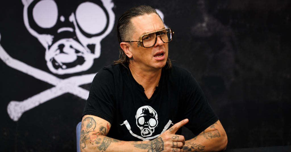 Close-up of Sid Wilson seated during an interview, wearing large tinted sunglasses, a black graphic T-shirt, and displaying tattooed arms while gesturing with one hand.