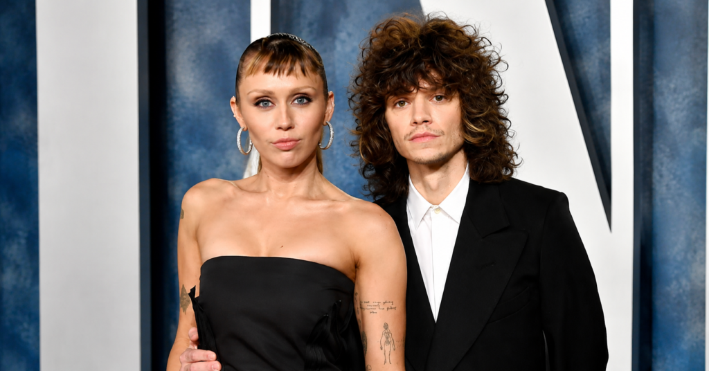 Maxx Morando stands beside Miley Cyrus on the red carpet wearing a black suit with a white shirt, while Miley wears a strapless black gown with hoop earrings and sleek bangs.