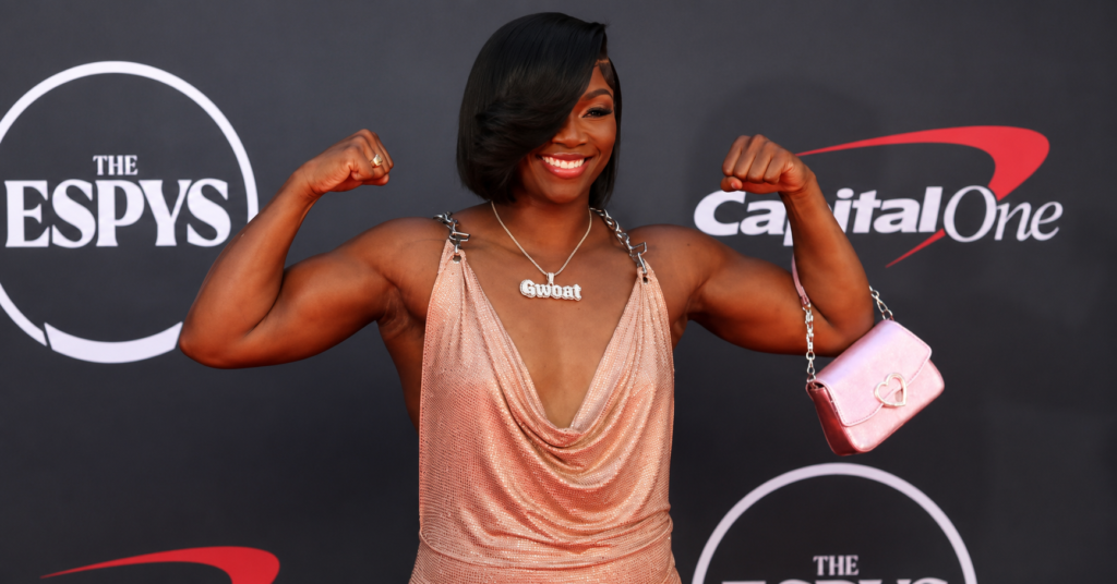 Claressa Shields flexes her arms while posing on the red carpet in a shimmering gown at a sports awards event.