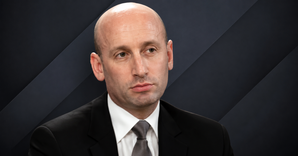 Close-up portrait of Stephen Miller wearing a black suit, white shirt, and gray tie against a dark background.