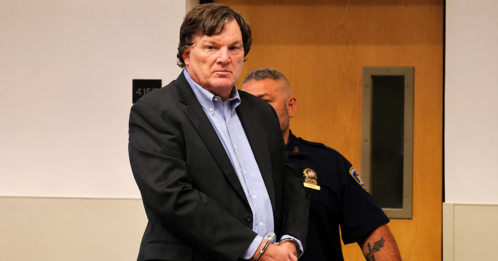 Rex Heuermann wearing a dark suit and light blue shirt, standing in handcuffs with a police officer behind him inside a courthouse.