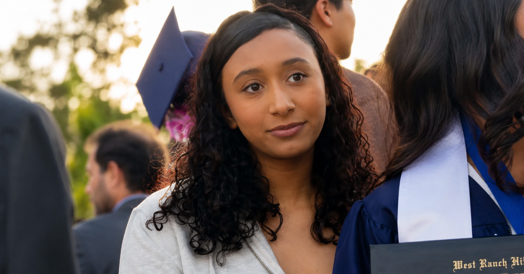 Celeste Rivas Hernandez wearing a light gray hoodie with curly dark hair, standing among graduates and guests at an outdoor graduation event.