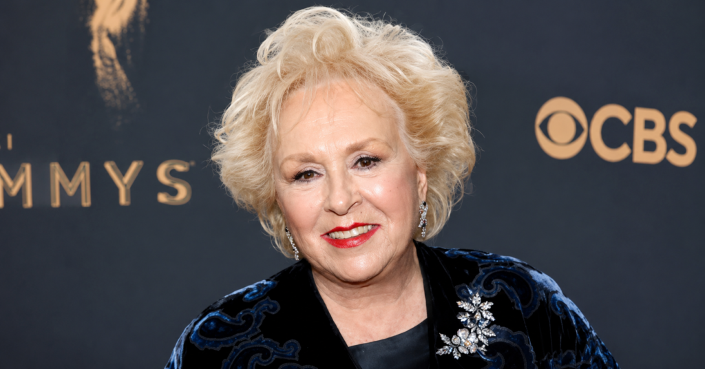 Portrait of Doris Roberts wearing a black velvet jacket with a jeweled brooch, red lipstick, and short blonde hair at a red carpet event.