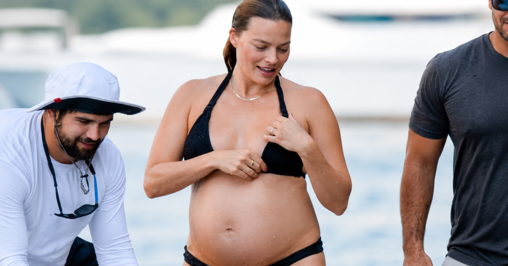 Margot Robbie, visibly pregnant, wearing a black bikini while walking near the beach with companions during a daytime outing.