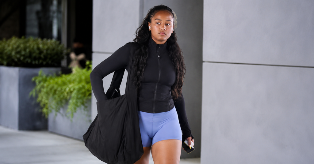 Sasha Obama walking in casual athletic wear with a black jacket, blue shorts, and a large black gym bag.