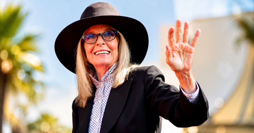 Portrait of actress Diane Keaton smiling and waving at a public event while wearing her signature wide-brim hat and black blazer.
