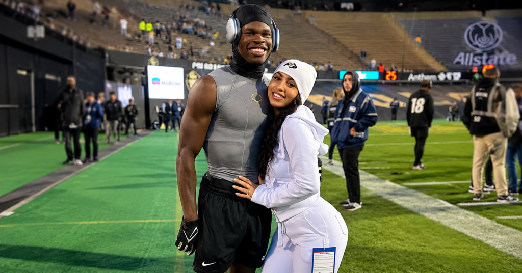 Leanna Lenee hugging Travis Hunter on the football field before a game while both smile for the camera in a stadium.