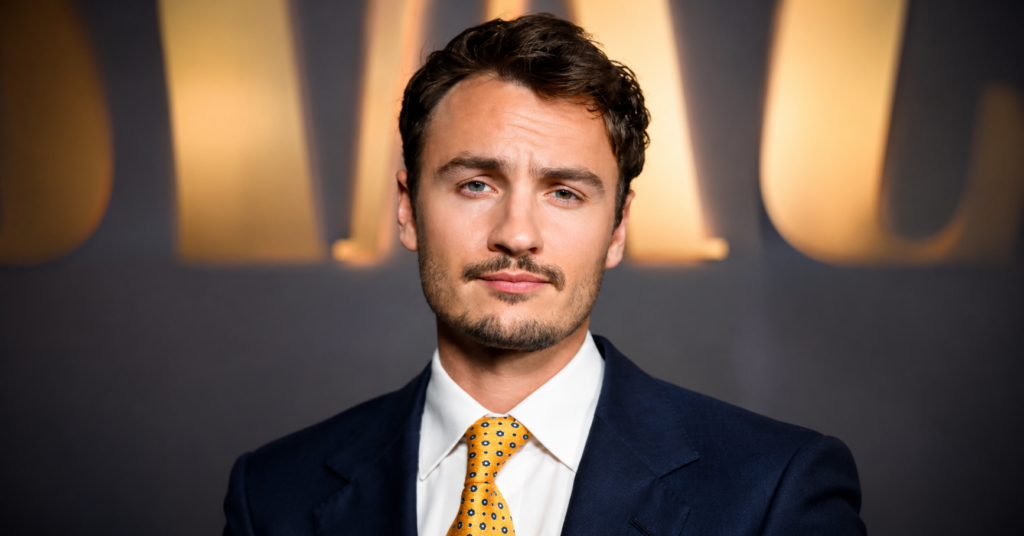 Brandon Thomas Lee posing for photographers at a red carpet event in a navy suit, white shirt, and gold patterned tie.