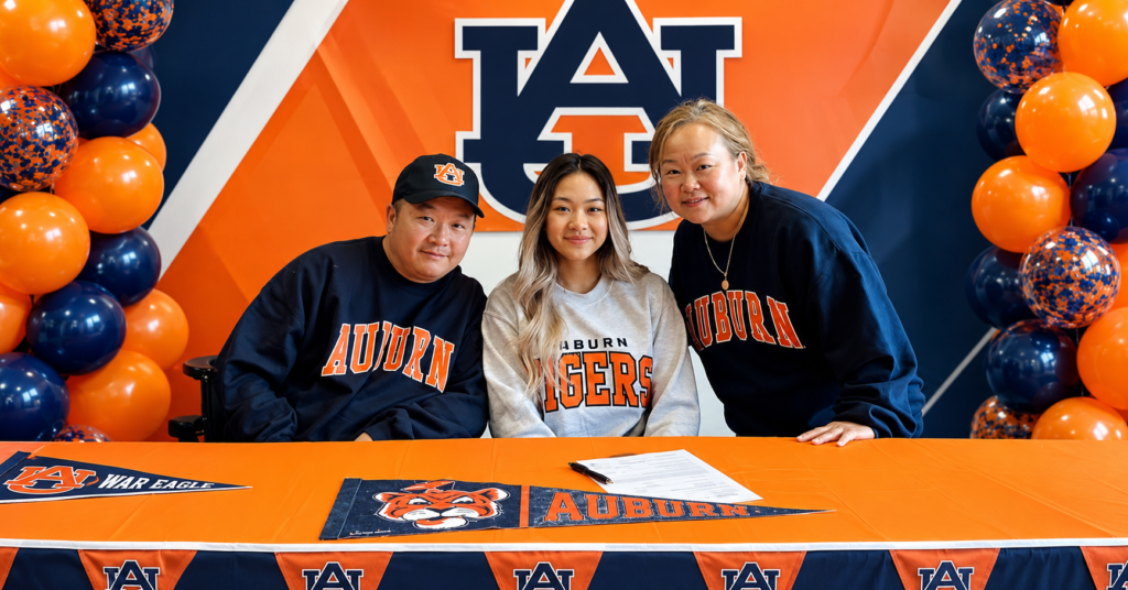 Suni Lee with her parents at Auburn University signing event, seated behind a table with Auburn decorations and an Auburn Tigers backdrop.