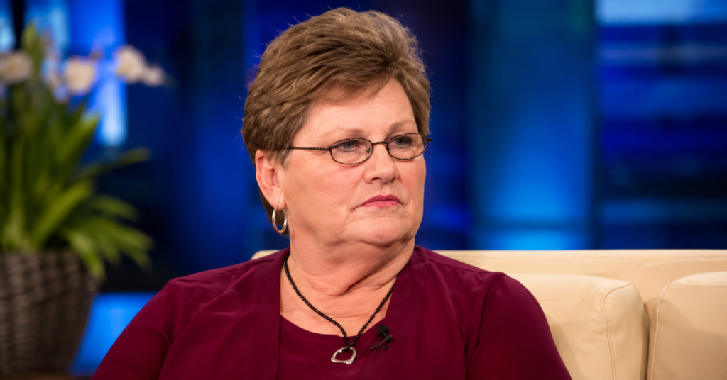 Christine Gacy wearing glasses and a burgundy top while seated on a television talk show set during an interview.