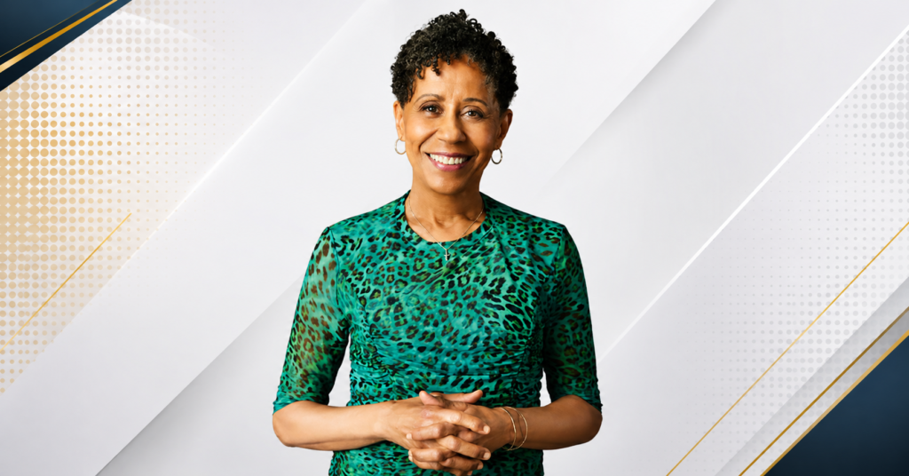Professional headshot of Vernee Watson smiling in a green animal-print blouse with hands clasped against a neutral gray background.