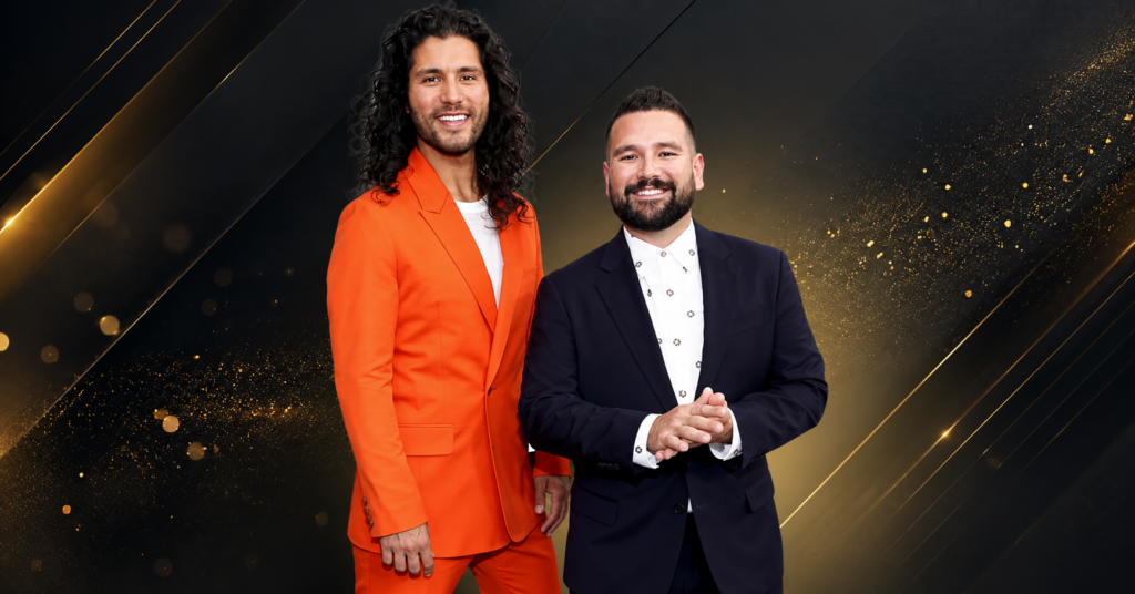 Dan Smyers and Shay Mooney pose together on the red carpet at a public event, with Shay wearing a dark suit and Dan wearing a bright orange suit.