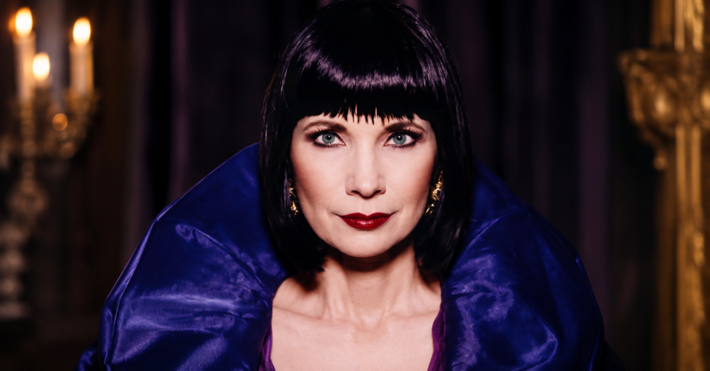 Studio portrait of Mystic Meg wearing a royal blue high-collar outfit with black bob hair, red lipstick, and a confident expression.