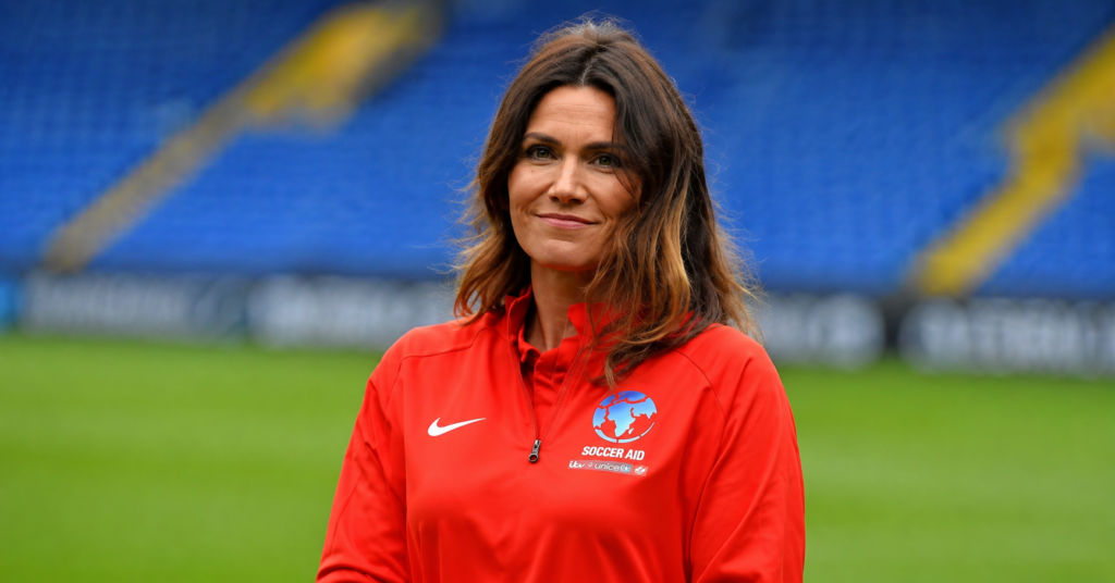 Susanna Reid standing on a football pitch in a red Soccer Aid Nike training jacket with stadium seating in the background.