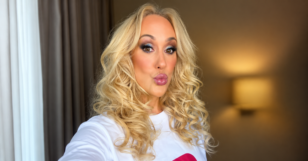 Brandi Love taking a close-up selfie with curled blonde hair, dramatic eye makeup, glossy lips, and a white top indoors.