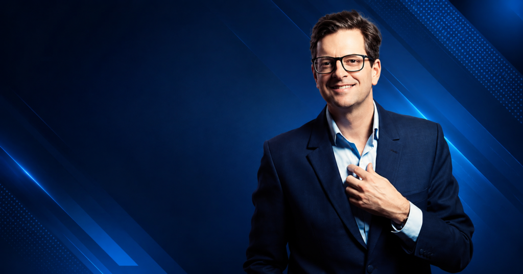 William Sitwell wearing a navy suit and glasses, smiling in a studio portrait with a neutral background.