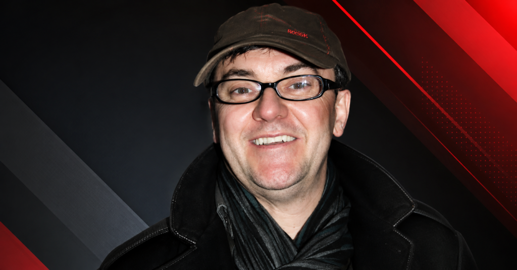Joe Pasquale, British comedian and television personality, smiling while wearing glasses, a dark cap, and a scarf.