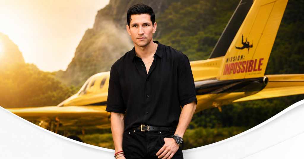 Danny Ramirez posing in a black shirt and black pants in front of a yellow airplane at a promotional event.