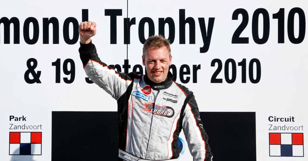 Jeroen Dik raises his fist in celebration while standing on the podium in a Porsche racing suit after a motorsport event victory.
