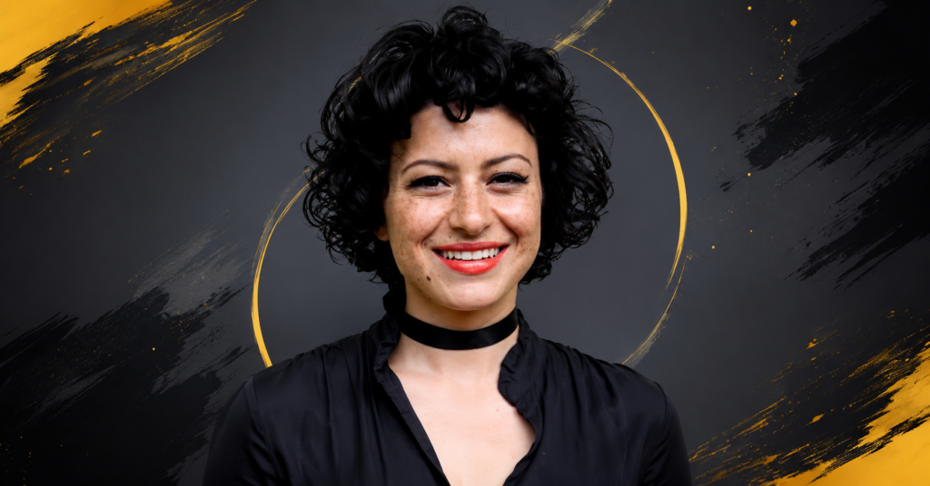 Alia Shawkat smiling in a black outfit with short curly hair at a red carpet event.