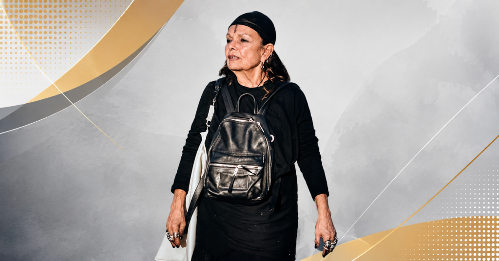 Michele Lamy wearing a black dress, headscarf, and leather backpack while walking outdoors at a public event.